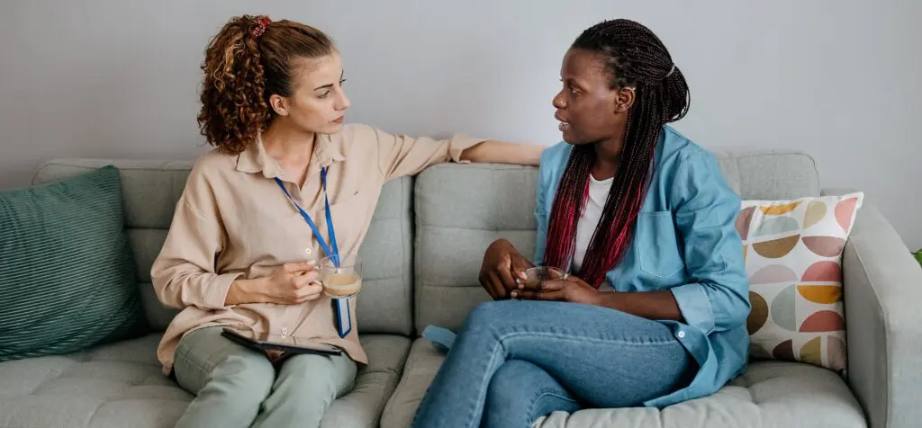 A social worker sits on a sofa holding a cup of tea speaking to a lady who is living with young onset dementia
